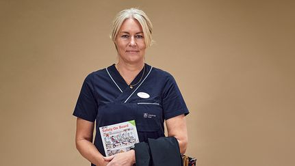 En person står mot beige bakgrund. Hon har på sig vårdkläder och håller ett säkerhetskort som finns på flygplan.