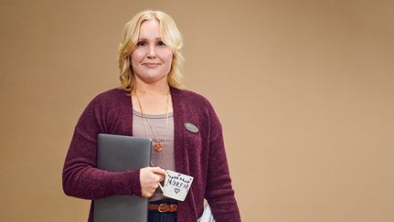 En person står mot beige bakgrund. Hon är vänd mot kameran och håller en laptop under ena armen och en kopp, där det står "Morfar" på.