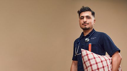 En person står mot beige vägg, han är vänd mot kameran och håller en kudde under armen.