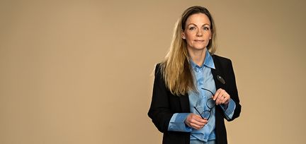 Helena står mot en beige bakgrund. Hon håller i ett par gladögon och titar in i kameran. Hon håller ett par glasögon i handen. Hon har en svart kavaj med blå skjorta under.