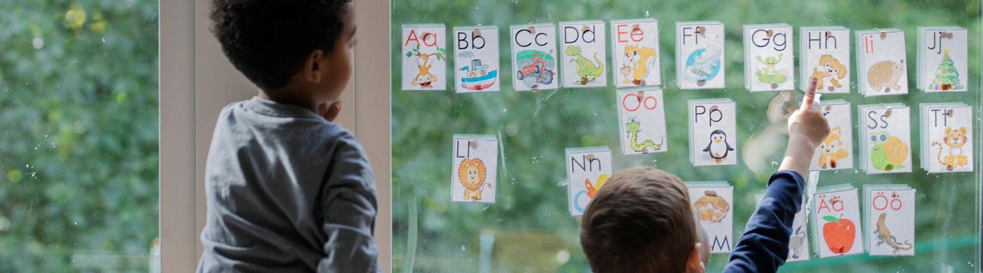Två barn står framför fönster med bilder på bokstäver inne i en förskolan. Den ena pojken pekr på en bokstav.