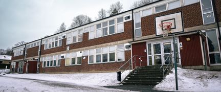 En entré till en skolbyggnad i rött tegel med en skolgård och en basketkorg framför.