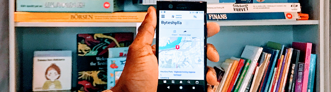 Hand håller upp mobil som visar Smarta Kartans hemsida framför Byteshyllan på Frilagret.