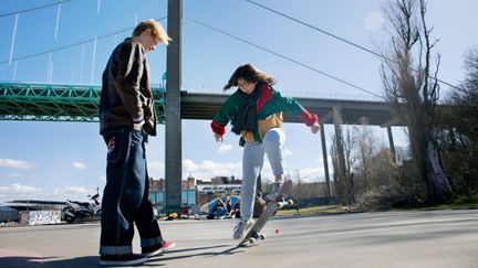 Två ungdomar utomhus. En tjej med skateboard och en kille som tittar på.