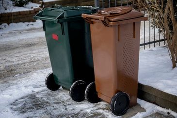 Ett grönt och en brunt avfallskärl ställda vid tomtgränsen till fastigheten. Det är skottat runt om kärlen och marken är frusen. 