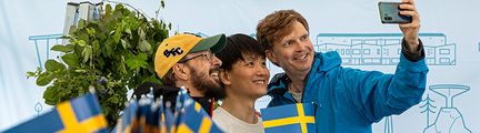 Tre personer tar en selfie framför en fotovägg. I förgrunden syns svenska flaggor.