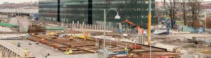 Byggarbetsplats med glasbyggnader i bakgrunden och en ofärdig konstruktion i förgrunden.