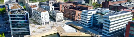 Bild på Lindholmshamnen en solig dag med ljusa byggnader vid hamnbassängen där det också finns ett stort bryggdäck med planteringar och sittplatser..