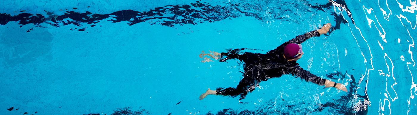 Person iklädd svart baddräkt och lila badmössa simmar genom blått vatten i en simhall.