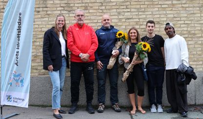 Framför en tegelvägg står sex personer. Några håller i blommor. 