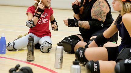 Flera unga personer sitter på ett sporthallsgolv iklädda rollerderbyutrustning.