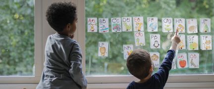 Två barn som står vid ett fönster och pekar på bokstäver som är upphängda på fönsterrutan