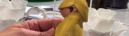 En hand håller en poslinsfigur med gula regnkläder. I bakgrunden finns olika porslinsskålar.
