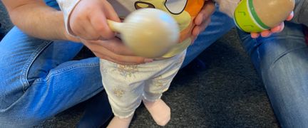 Ett barn håller i skallror och står framför en vuxen som sitter på golvet.