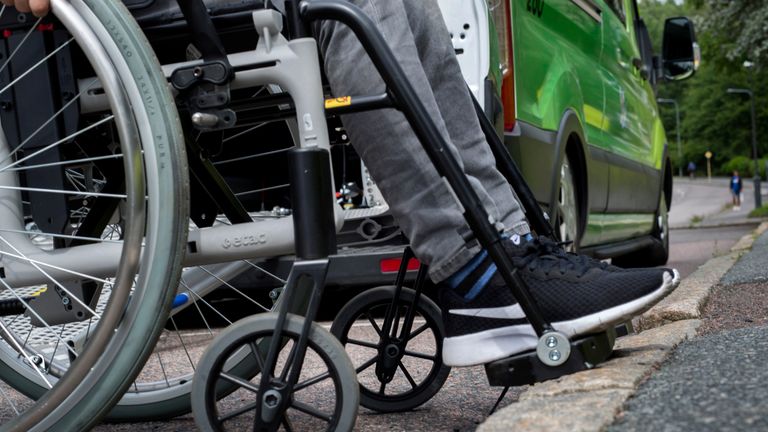 Närbild på rullstolshjul och ben på fotstödet på rullstolen. I bakgrunden syns nedre kanten av en grön färdtjänstbuss.
