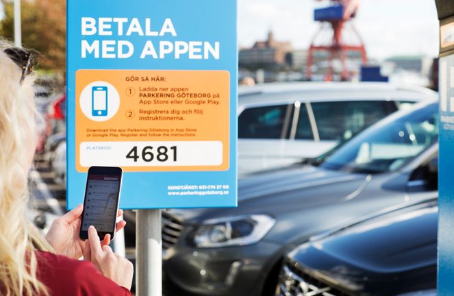 En kvinna använder en app i mobilen för att betala parkeringsavgift. Mobiltelefonen hålls framför en skylt bredvid parkerade bilar.