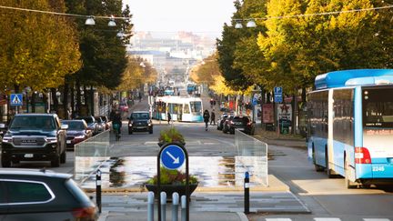 Götaplatsen med vattenspelsfontän i mitten av gatan. På ena sidan kör flera bilar i tät rad mot kameran tillsammans med en cyklist. På andra sidan gatan står en buss vid sin hållplats.