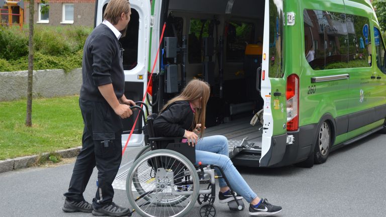 En man kör en tjej i rullstol mot en större grön färdtjänstbil som har en utrullad ramp.