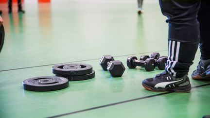 Ett par fötter syns på ett grönt golv. Bredvid ligger viktplattor och hantlar.