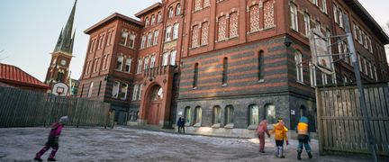 En grupp barn som går på skolgården framför en en stor skolbyggnad i rött tegel.