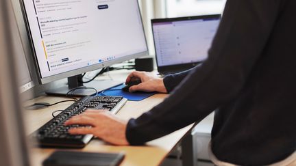 En person står och jobbar vid sitt tangentbord och skärm på ett skrivbord.
