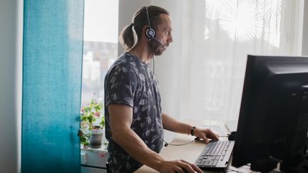 En person med headset som står vid ett skrivbord och jobbar vid en dator. 
