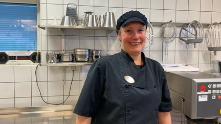 En person i måltidskläder står i ett kök och tittar glatt in i kameran.
