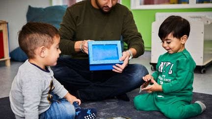 Två barn sitter på golvet. En pedagog visar upp en skylt.