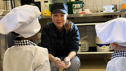 En person i kockkläder sitter på huk framför två barn i kockkläder och pratar med dem.