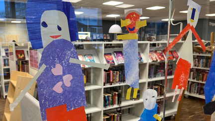 Barns målningar av personer sitter uppsatta på en glasvägg i biblioteksmiljö.