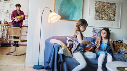 Förälder med spädbarn i bakgrunden och två äldre barn som leker i förgrunden.. i