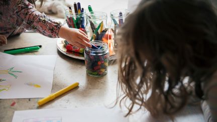 Två barn sitter tillsammans och målar. Ena barnet sträcker sig efter kritor i en burk.