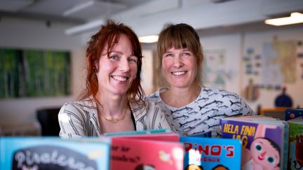 Två personer står framför barnböcker som är uppradade framför dem. De ler och tittar in i kameran.