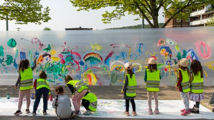 Flera barn i reflexvästar står och målar regnbågar på en stor uppspänd målarduk.