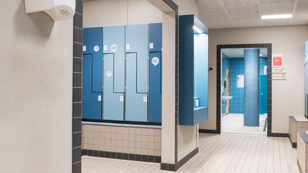 Omklädningsrum med skåp i olika blå nyanser och vitt kakel på golvet.