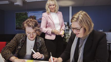 Två kollegor jobbar tillsammans vid ett bord. En mötesledaren övervakar samtalet från sidan a