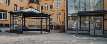 En paviljong med en bänk i framför en gul skolbyggnad med en entré
