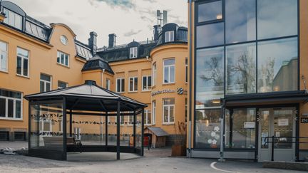 En paviljong med en bänk i framför en gul skolbyggnad med en entré