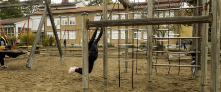 En elev som hänger i en lekställning i sanden på en skolgård med en skolbyggnad i bakgrunden.