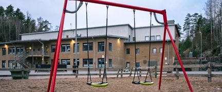 En röd gungställning i en sandlåda framför en skolbyggnad i gult tegel.
