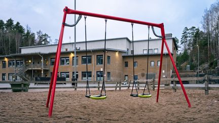 En röd gungställning i en sandlåda framför en skolbyggnad i gult tegel.