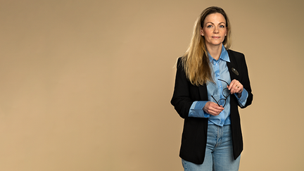 Helena står mot en beige bakgrund. Hon håller i ett par gladögon och titar in i kameran. Hon håller ett par glasögon i handen. Hon har en svart kavaj med blå skjorta under.