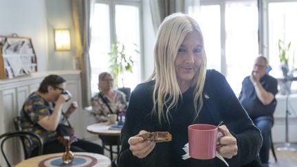 Ett foto av en person som står med en tekopp och kaka i händerna i en kafé-miljö.