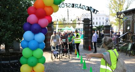 I förgrunden en portal av färgglada ballonger. I bakgrunden kommer personer med rullatorer gående.