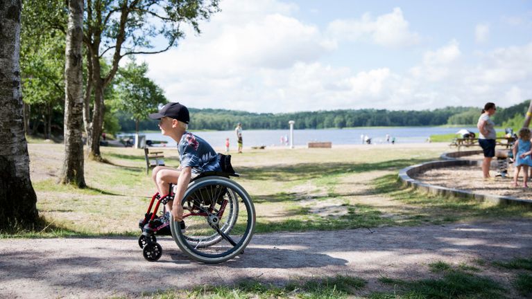 Ung kille rullar rullstol vid en strand vid en sjö