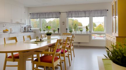 Allrum med långbord och köksdel. Genom fönstren syns grönska.
