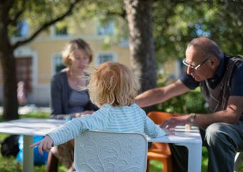En yngre kvinna, en äldre man och ett barn med ryggen mot kameran sitter tillsammans utomhus runt ett bord.
