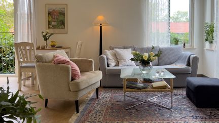  Ett vardagsrum med soffa, fåtölj och glasbord med en vas blommor. I bakgrunden syns ett matbord, en golvlampa och tavla på väggen.