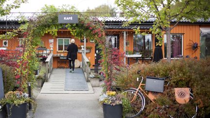 En svartklädd person är går mot ett orange trähus omgivet av träd, buskar och blommor. En cykel står vid infarten.