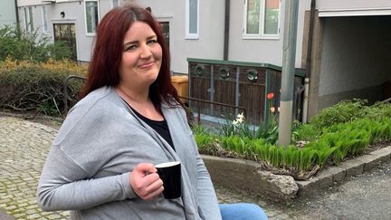 En person sitter på innergården framför ett grått trähus med en kaffekopp i handen.